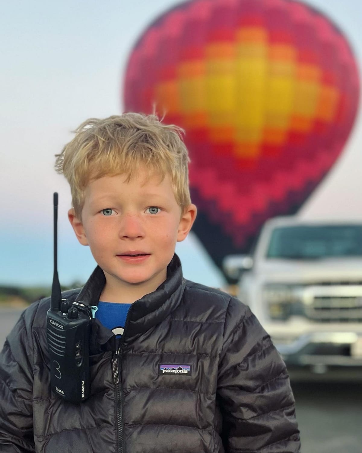 Young Boy with Walkie-Talkie in Front of Colorful Hot Air Balloon and White Truck at Sunrise Event