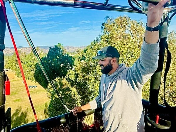 Man Enjoying Scenic Hot Air Balloon Ride Above Lush Greenery with Distant Mountain Views on a Clear Day