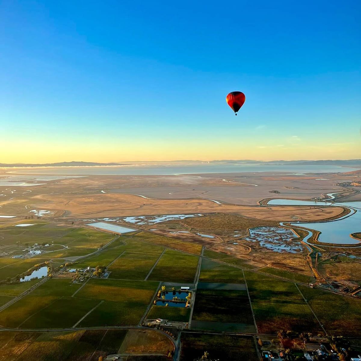 Solitary Red Hot Air Balloon Floating Over Expansive Patchwork of Fields and Winding River at Sunrise with Clear Blue Sky