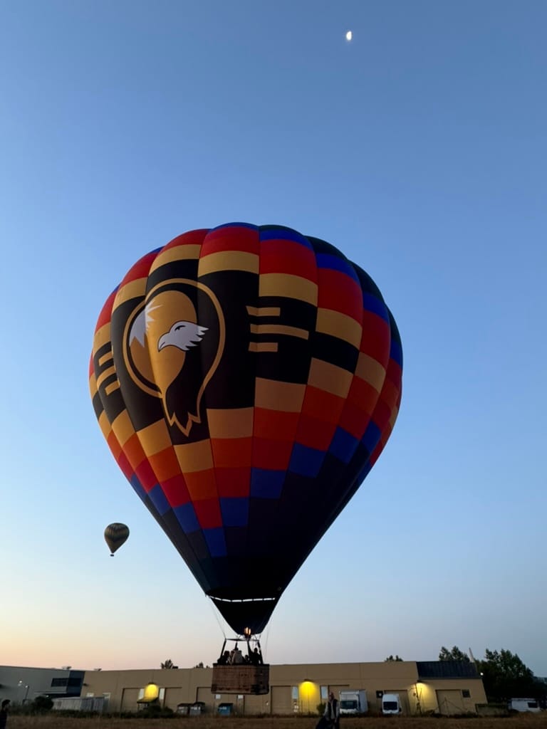 Colorful Hot Air Balloon with Eagle Emblem Ascends Against a Clear Evening Sky with Moon Over Industrial Buildings