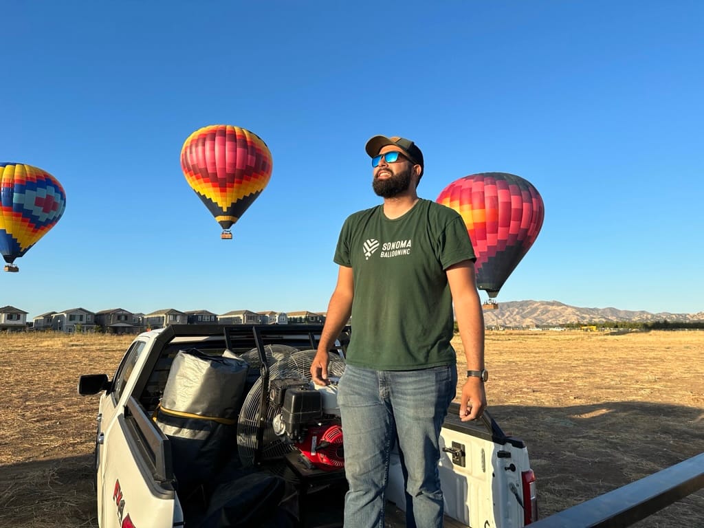 Man Standing on Truck Bed Against Clear Blue Sky with Colorful Hot Air Balloons in the Background and Open Field Landscape
