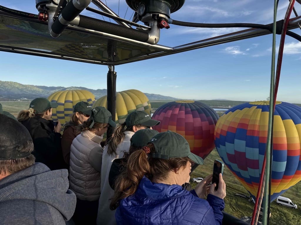 Group of People in a Hot Air Balloon Basket Capturing Scenic Views of Vibrant Multicolored Balloons Against a Backdrop of Rolling Hills and Blue Sky
