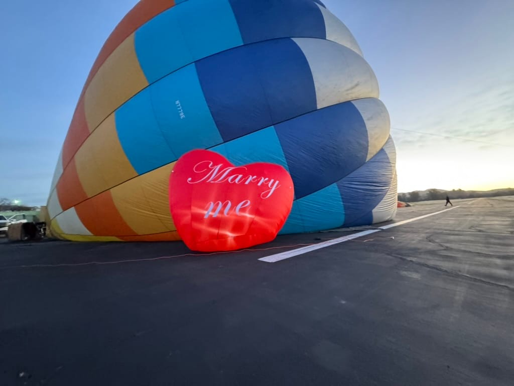 Deflated Multicolored Hot Air Balloon with Romantic 'Marry Me' Red Heart Sign at Dusk on an Open Airfield