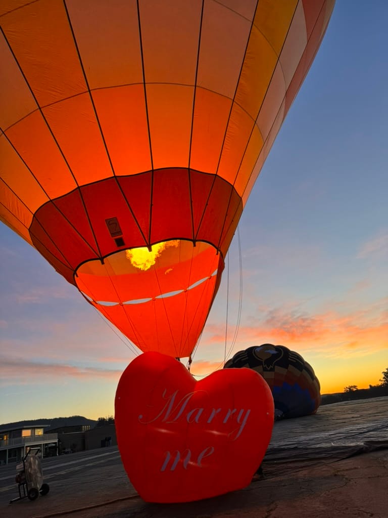 Romantic Hot Air Balloon Proposal at Sunset with Illuminated Heart-Shaped 'Marry Me' Sign and Vibrant Sky Background