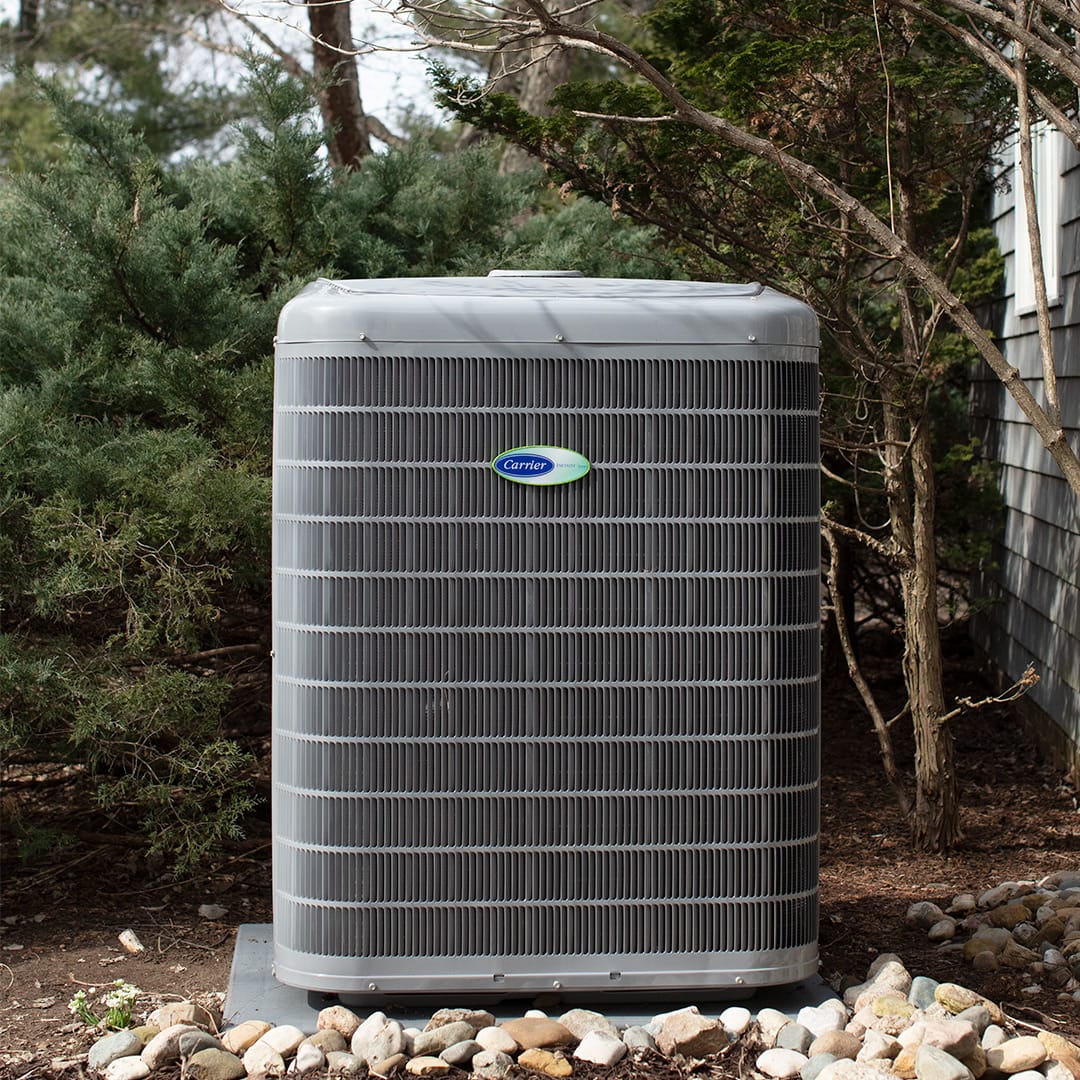 Outdoor Air Conditioning Unit with Metal Grille Surrounded by Greenery and Stones Near a Building Exterior