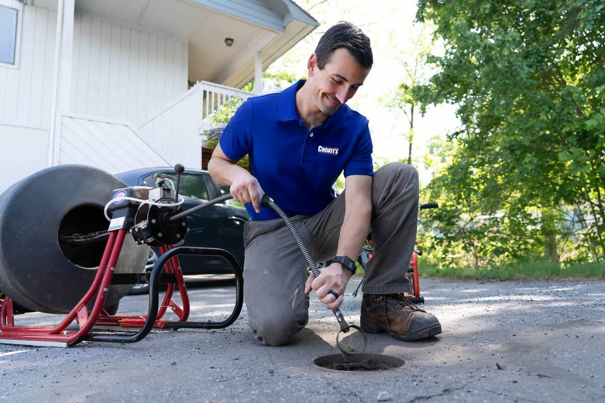 Plumber in Blue Uniform Using Drain Snake to Clear Outdoor Pipe Blockage Near Residential Building