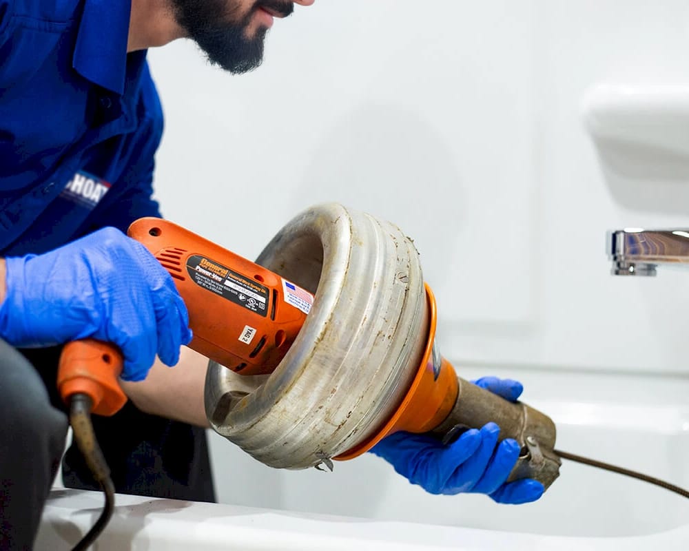 Professional Plumber Utilizing an Electric Drain Snake to Unclog Bathtub Drain, Wearing Blue Gloves and Uniform in a Bathroom Setting