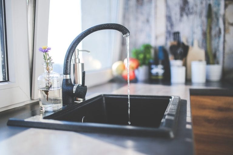 Modern Kitchen Sink with Running Water and Decorative Elements Including a Small Vase with Flowers on a Sunlit Countertop Near a Window with a View of Cozy Kitchen Essentials in the Background