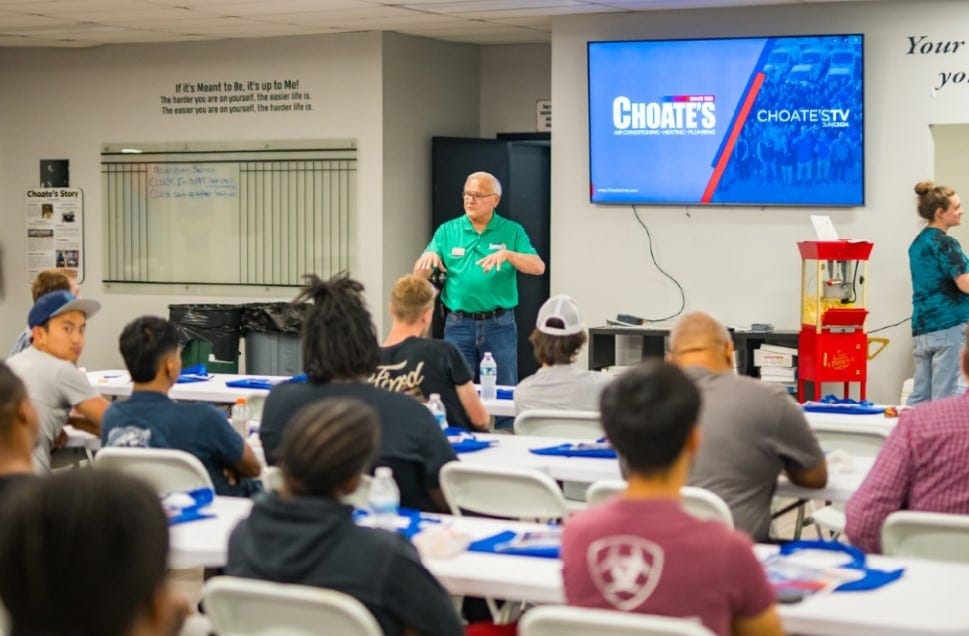 Training Session in Classroom with Instructor Speaking to Attendees Seated at Tables, Large Screen Displaying Choate's Branding, and Popcorn Machine in Background