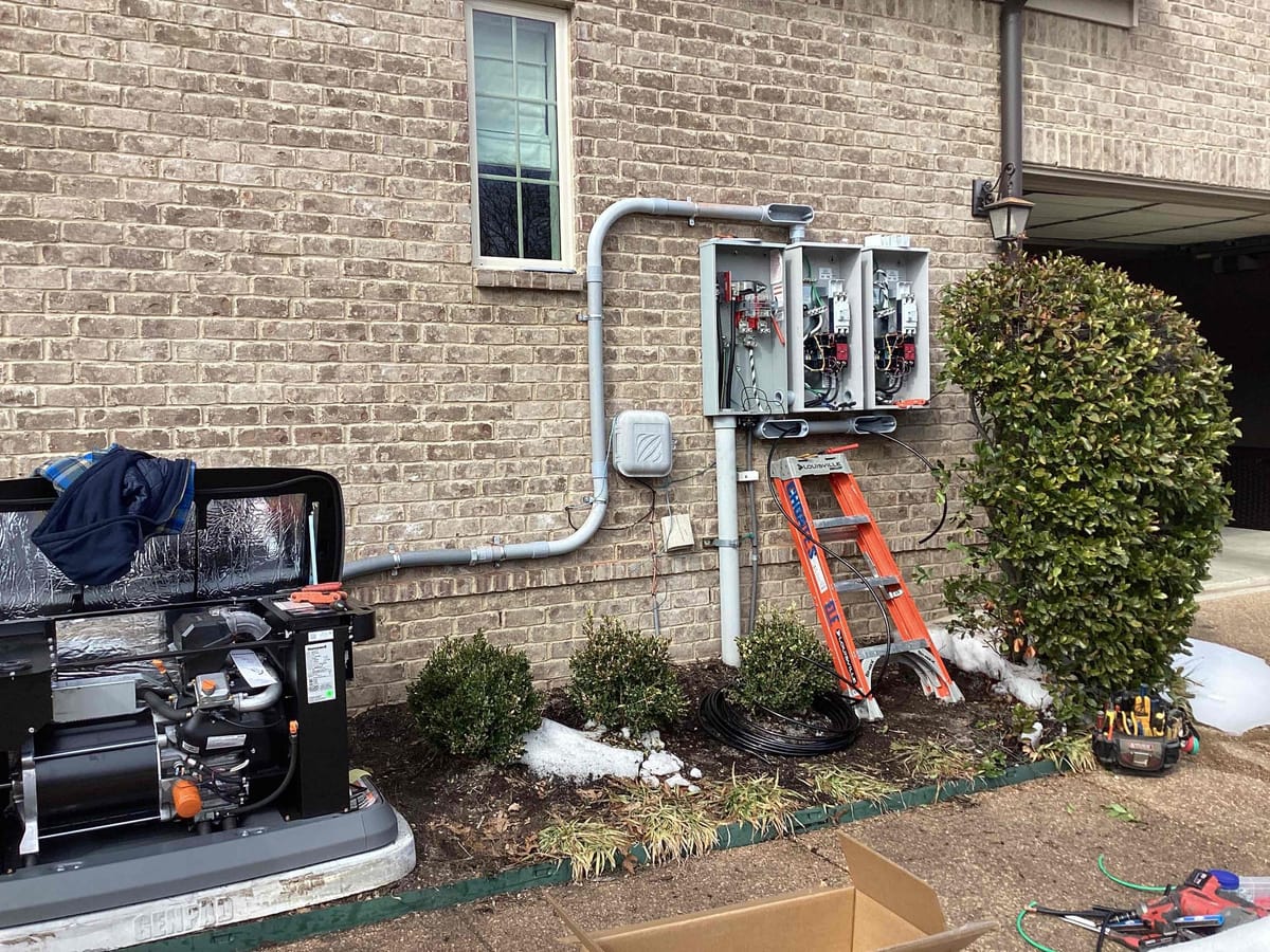 Outdoor Generator Installation with Open Electrical Panels and Tools on Brick Wall Exterior Near Garage and Shrubbery