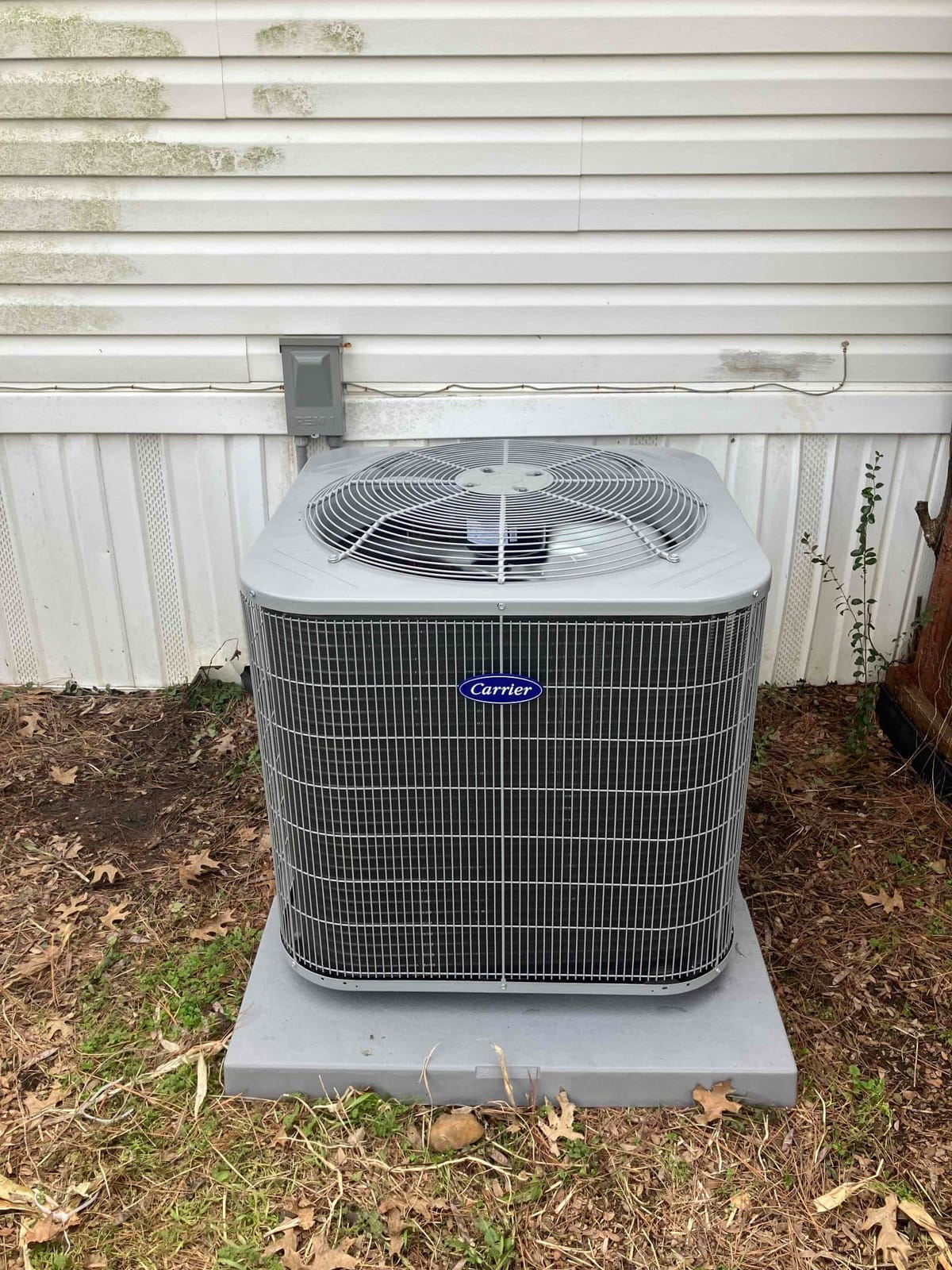 Outdoor Air Conditioning Unit with Protective Grille Installed on Concrete Slab Next to White Vinyl-Sided Building with Visible Weathering and Ground Covered in Dry Leaves and Grass.