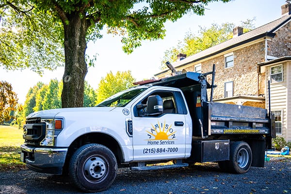 White Home Services Truck Parked on Gravel Driveway Beside Stone House with Lush Greenery and Tree in Background