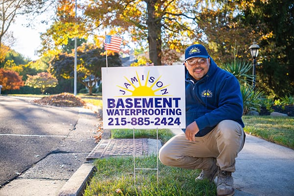 Smiling Man in Blue Hoodie Kneeling Beside 'Jamison Basement Waterproofing' Sign on Suburban Sidewalk with Autumn Foliage and American Flag in Background