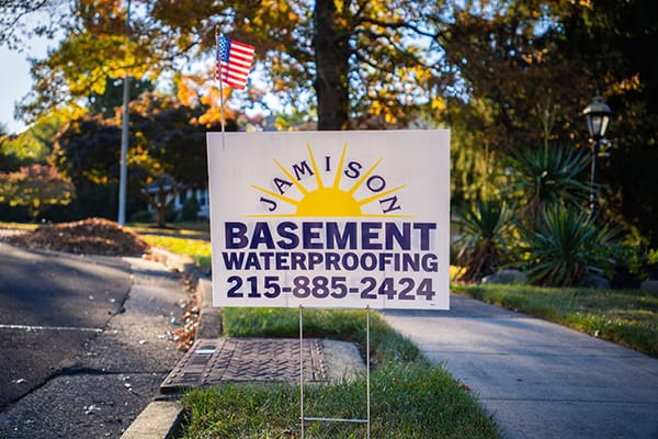 Lawn Sign Advertising Basement Waterproofing Service with Contact Number, Displayed on a Sunny Residential Street with Autumn Foliage and Small American Flag
