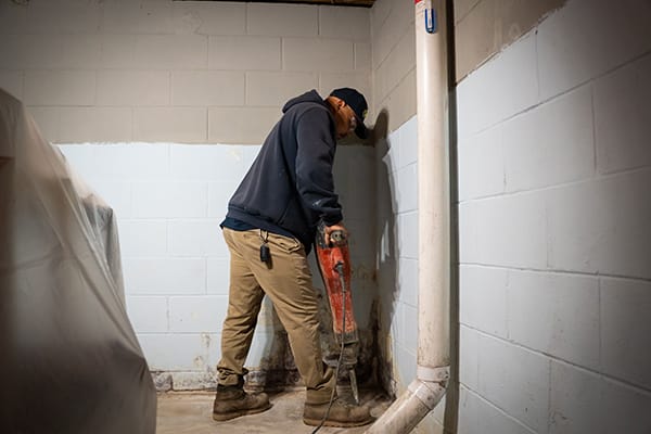 Worker in Basement Using Jackhammer on Concrete Floor Near Cinder Block Wall and PVC Pipe