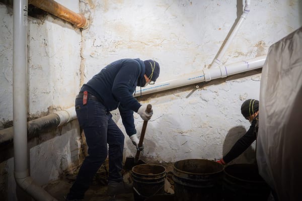 Two Workers in Protective Gear Digging and Removing Soil in a Dimly Lit Basement with Exposed Pipes and Buckets
