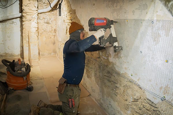 Worker in Protective Gear Drilling Mesh Barrier onto Old Stone Basement Wall with Industrial Equipment