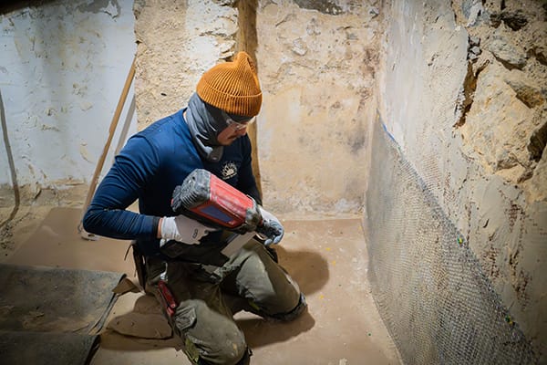 Worker in Protective Gear Using Power Tool to Secure Metal Mesh on Basement Wall with Exposed Stones and Worn Plaster