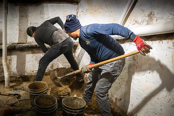 Workers Engaged in Manual Labor Digging Dirt in a Dimly Lit Basement with Buckets and Pipes Nearby